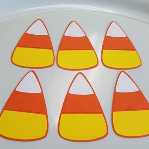 May include: Six paper candy corn cutouts in orange, yellow, and white. The cutouts are in the shape of candy corn and are perfect for crafts or decorations.