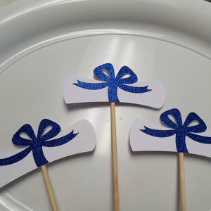 May include: Three graduation cap cupcake toppers with blue glitter bows and white scrolls. The toppers are on wooden sticks.
