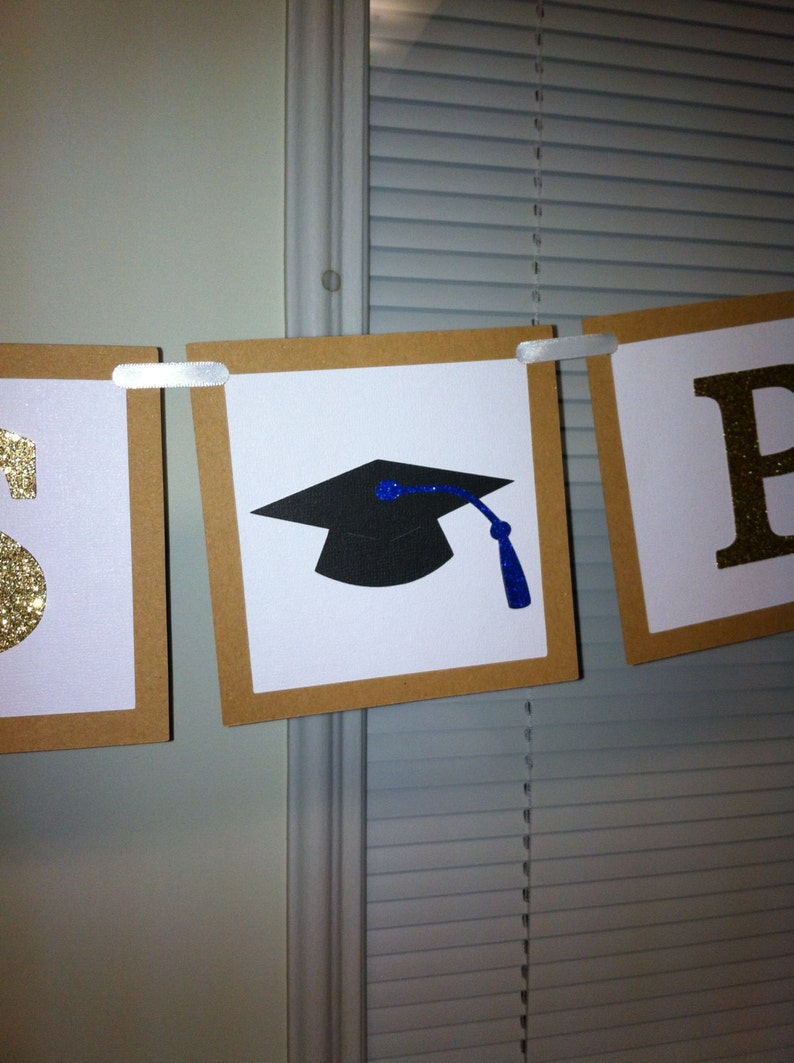 May include: A graduation banner with gold glitter letters that spell "Class of" and a black graduation cap with a blue tassel. The banner is made of brown cardstock and white paper.