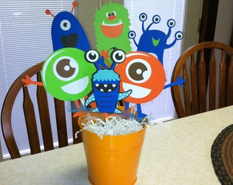 May include: A centerpiece featuring a variety of colorful monster characters made of paper. The monsters are arranged in a small orange bucket with white shredded paper.