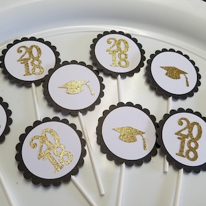 May include: Black and white cupcake toppers with gold glitter accents. The toppers feature the year 2018 and a graduation cap.