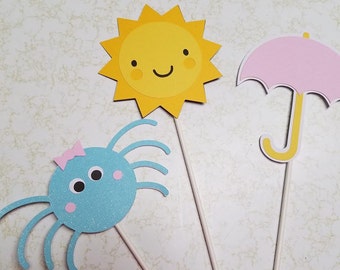 May include: Three paper craft decorations on sticks. A yellow sun with a smiling face, a pink umbrella, and a blue spider with a pink bow.