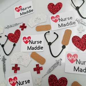 May include: A collection of red and white confetti with a heart beat symbol and the text "Nurse Maddie". The confetti includes red glitter hearts, white glitter crosses, white glitter nurse hats, black and white stethoscope outlines, silver syringes, and brown bandaids.