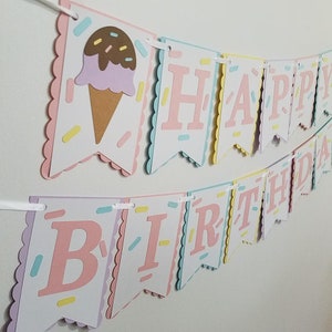 May include: A pink, white, and blue Happy Birthday banner with an ice cream cone on the first flag. The banner has a scalloped edge and is decorated with sprinkles.