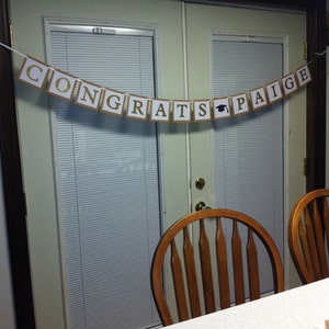 May include: A brown and gold banner with the text "Congrats Paige" hangs on a white door with a wood frame. The banner has a graduation cap icon between the words "grats" and "Paige".