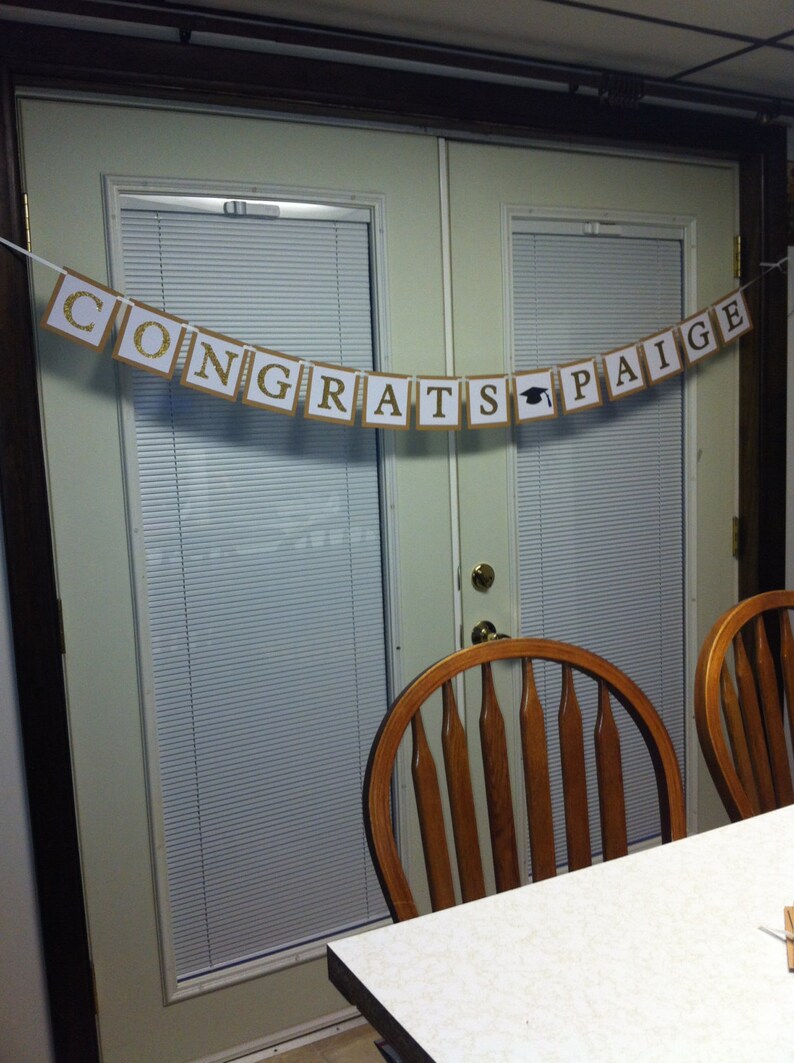 May include: A brown and gold banner with the text "Congrats Paige" hangs on a white door with a wood frame. The banner has a graduation cap icon between the words "grats" and "Paige".