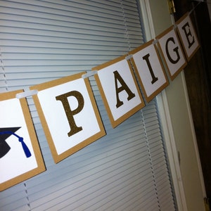 May include: A graduation banner with the name "PAIGE" in gold glitter letters on white squares. The banner is strung with white ribbon and has a graduation cap on the left side.
