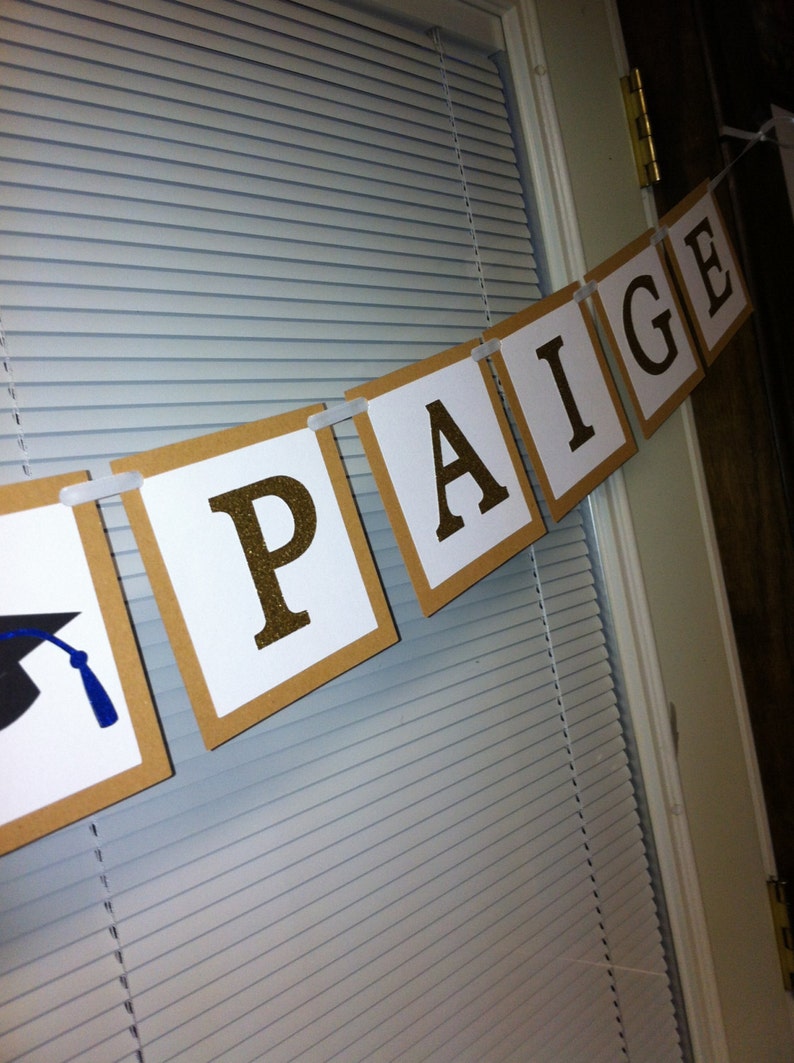 May include: A graduation banner with the name "PAIGE" in gold glitter letters on white squares. The banner is strung with white ribbon and has a graduation cap on the left side.