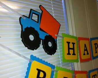 May include: A blue and orange construction truck with black wheels hangs from a white string banner. The banner reads "Happy Birthday" in yellow letters with green, red, and blue borders.