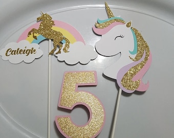 May include: Three cake toppers featuring a unicorn, a rainbow, and the number 5. The unicorn is white with pink, purple, and blue glitter. The rainbow is yellow, pink, and blue. The number 5 is pink with gold glitter. The cake toppers are on white sticks. The name "Caleigh" is written on a white cloud.