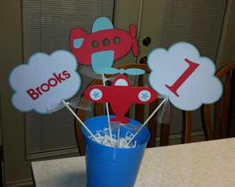 May include: A blue bucket centerpiece with white shredded paper inside. There are red and blue paper airplanes and clouds with the words "Brooks" and "1" on them.