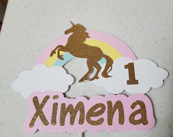 May include: A pink and yellow rainbow with a gold glitter unicorn jumping over it. There are two white clouds and a pink banner with the name "Ximena" and the number "1" in gold glitter.