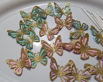 May include: A collection of 12 paper butterfly cake toppers in pink, blue, and gold glitter. The butterflies are layered and have a delicate, intricate design.