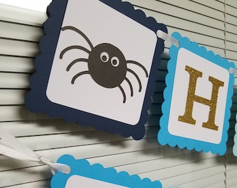 May include: A blue and white banner with a spider and the letter H in gold glitter. The banner is decorated with scalloped edges.