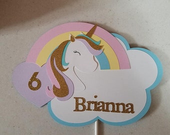May include: A white unicorn with a rainbow and a purple heart with the number 6 on it. The unicorn is on a blue and white cloud with the name Brianna written in gold glitter.