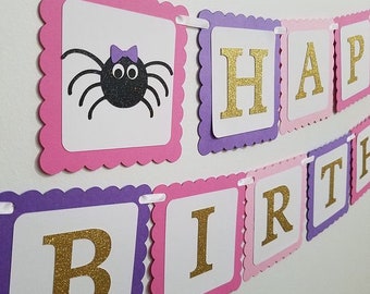 May include: A pink and purple banner with the words "Happy Birthday" in gold glitter letters. The banner also features a black spider with a purple bow on a pink square.