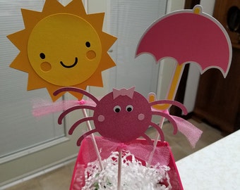 May include: A pink flower pot with white shredded paper inside. There are three paper decorations in the pot: a yellow sun with a smiling face, a pink spider with a bow, and a pink umbrella.