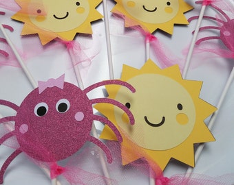 May include: A set of six cake toppers featuring pink spiders and yellow suns with happy faces. The toppers are attached to white sticks and have pink tulle bows.