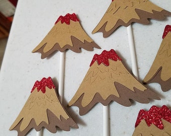 May include: Six brown paper cupcake toppers shaped like volcanoes with red glitter on the top. The toppers are on white sticks.