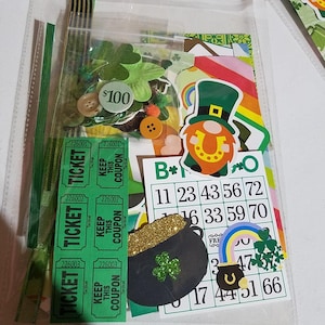 May include: A clear plastic bag filled with green, gold, and rainbow-colored craft supplies for a St. Patrick's Day themed project. The bag includes shamrocks, a pot of gold, a leprechaun, tickets, and a bingo card with numbers and a shamrock.