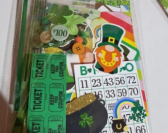 May include: A clear plastic bag filled with green, gold, and rainbow-colored craft supplies for a St. Patrick's Day themed project. The bag includes shamrocks, a pot of gold, a leprechaun, tickets, and a bingo card with numbers and a shamrock.