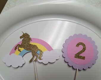 May include: Two cake toppers, one with a gold glitter unicorn silhouette on a rainbow and clouds, and the other with a pink circle with a gold glitter number 2.