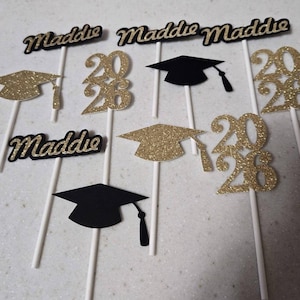 May include: Graduation cake toppers with the name "Maddie" in gold glitter script, along with gold glitter mortarboards and the numbers "2026". Some toppers are black mortarboards. All are attached to white sticks.