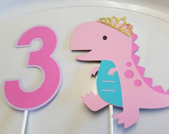 Puede incluir: Topper de pastel de dinosaurio rosa con una corona dorada y una barriga turquesa. El dinosaurio sostiene un número tres rosa.