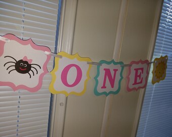 May include: A pink, yellow, blue and white banner with the word "ONE" in pink letters. The banner has a spider with a pink bow on the left side and a yellow sun on the right side.