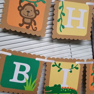 May include: A brown and tan paper banner with scalloped edges. The banner spells out "Happy Birthday" with each letter on a separate square. The letters are white and the squares have different designs. One square has a monkey hanging from a vine, another has a green alligator, and another has green grass.