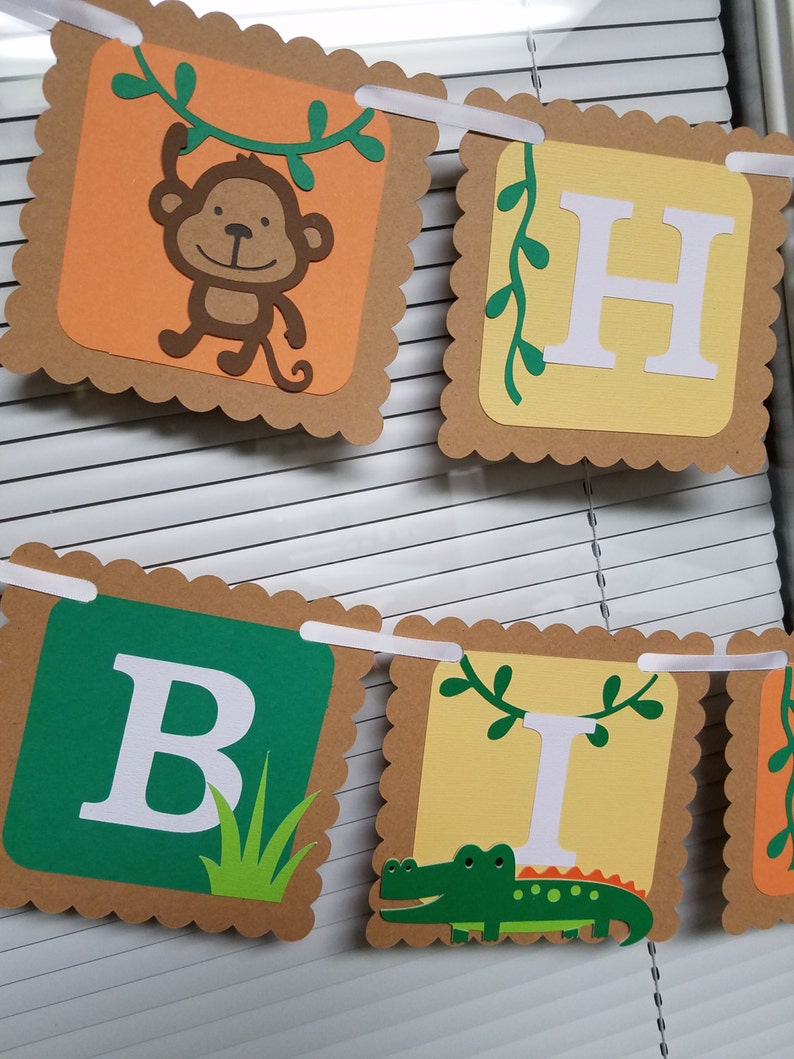 May include: A brown and tan paper banner with scalloped edges. The banner spells out "Happy Birthday" with each letter on a separate square. The letters are white and the squares have different designs. One square has a monkey hanging from a vine, another has a green alligator, and another has green grass.