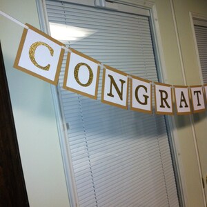 May include: A gold glitter banner with the word "CONGRATULATIONS" on it. The banner is hanging on a white door with blinds.