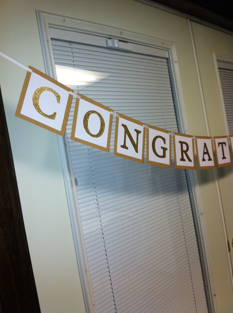 May include: A gold glitter banner with the word "CONGRATULATIONS" on it. The banner is hanging on a white door with blinds.