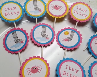 May include: Set of cupcake toppers featuring a yellow sun, a pink spider, and a blue pipe with water drops. The toppers have a scalloped edge and are printed with the words "Itsy Bitsy".