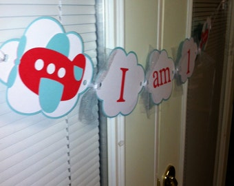 May include: A white and red banner with the words "I am 1" on a white door. The banner is decorated with blue and red airplanes and white tulle.