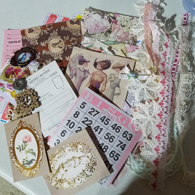 May include: A pink and white bingo card with numbers 5, 8, 22, 27, 41, 45, 46, 56, 63, 74, 48, 65, and 1. The card is surrounded by various craft supplies, including lace, ribbon, and paper scraps.