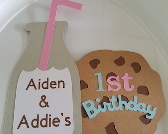 May include: A brown paper cutout of a cookie with the text "1st Birthday" in blue and pink letters. A brown paper cutout of a milk bottle with a pink straw and the text "Aiden & Addie's" in brown letters.