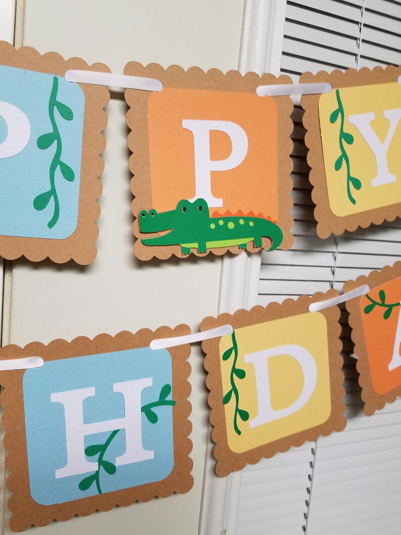 May include: A Happy Birthday banner with brown scalloped edges and white letters. The letters are on different colored squares: blue, orange, yellow, and light blue. Each square has a green vine with leaves. The letter P has a green alligator on it.