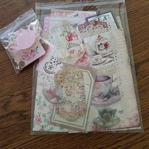 May include: A clear plastic bag filled with various tea-themed paper ephemera, including teacups, teapots, and floral designs. The bag also contains a small pink teapot and lace embellishments.