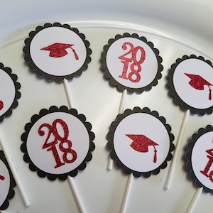 May include: A collection of graduation cupcake toppers. Each topper features a white circle with a black scalloped border. Some have the number "2018" in red glitter, while others display a red glitter graduation cap.