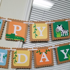 May include: A brown and orange Happy Birthday banner with a green alligator, a zebra, and green vines. The letters are white and the banner is strung together with white ribbon.