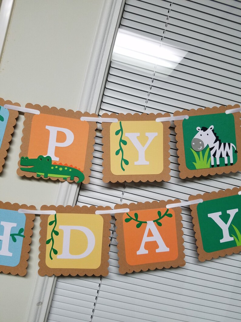 May include: A brown and orange Happy Birthday banner with a green alligator, a zebra, and green vines. The letters are white and the banner is strung together with white ribbon.