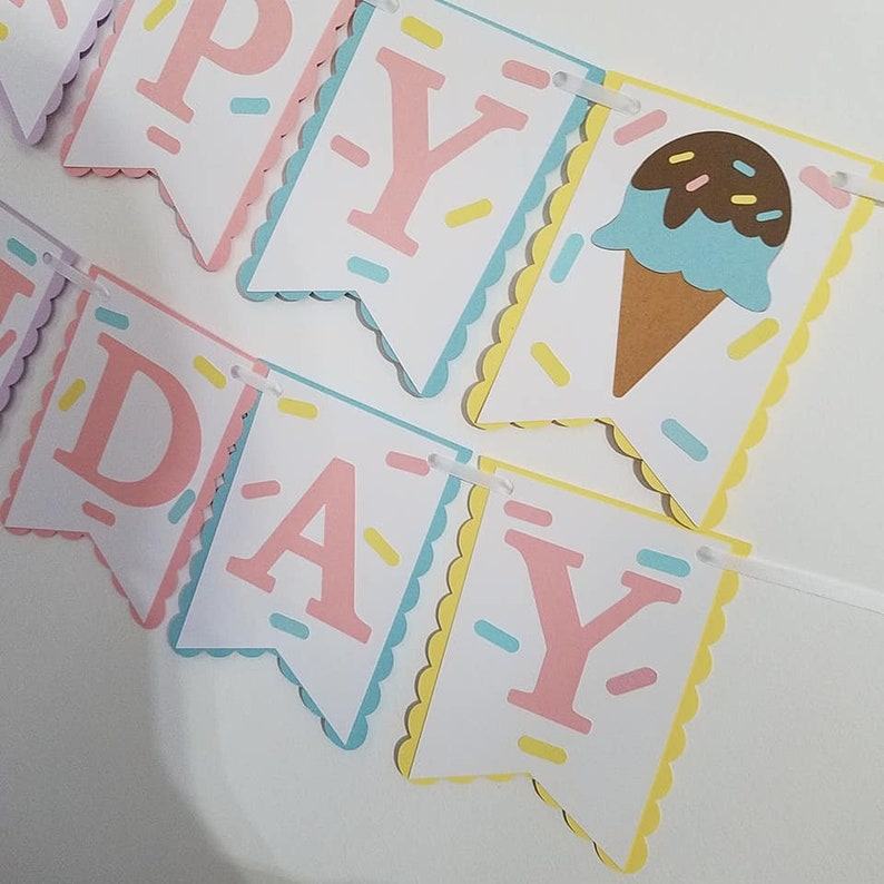 May include: A colorful Happy Birthday banner with pink letters and an ice cream cone graphic. The banner is made of paper and has a scalloped edge.