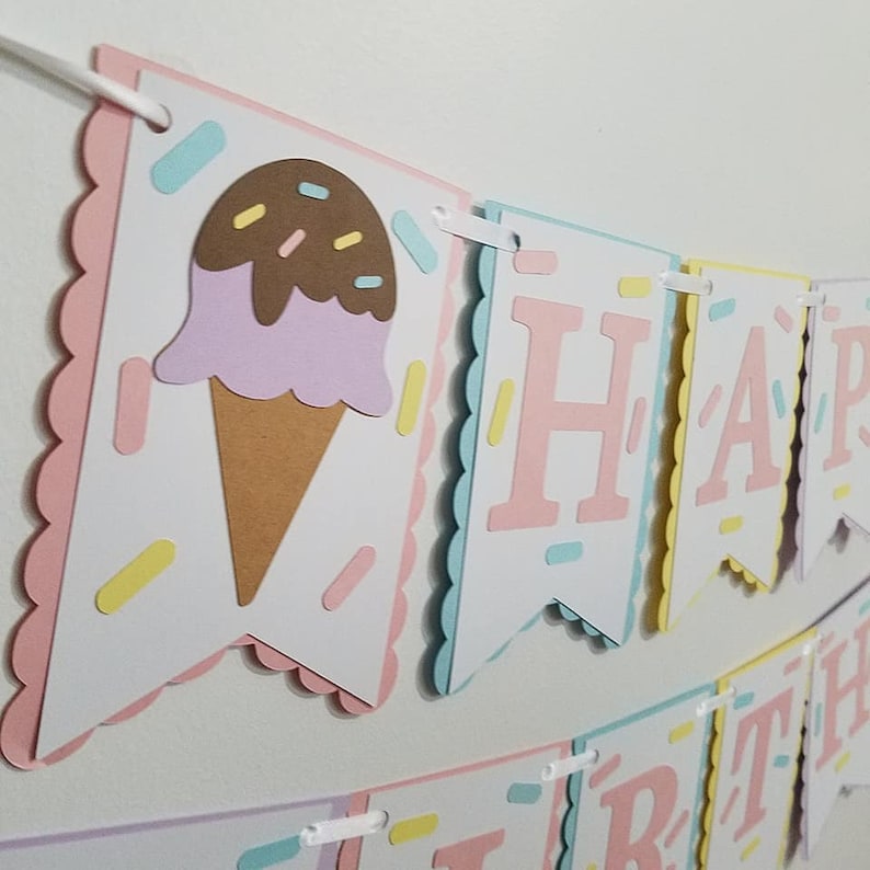 May include: A pink, blue, and yellow Happy Birthday banner with an ice cream cone on the first flag. The banner is made of cardstock and has a scalloped edge.