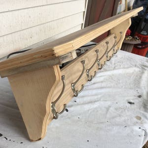 May include: A wooden wall shelf with a light finish, featuring a row of metal hooks. The shelf has a simple design with a flat top and a decorative lower edge. The hooks are evenly spaced along the bottom.