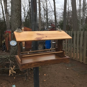 May include: A wooden bird feeder with a pitched roof, mounted on a metal pole. The feeder has a rectangular base and a tray for food. The wood is stained a medium brown color. The background includes trees and a fence.