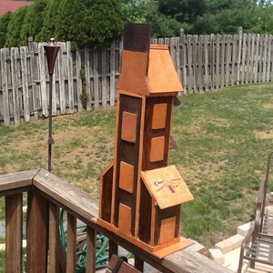 May include: A wooden birdhouse shaped like a tower with a brown and orange paint finish. The birdhouse has multiple compartments and a dragonfly design on the front.