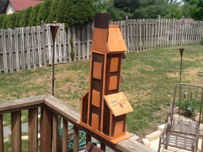 May include: A wooden birdhouse shaped like a tower with a brown and orange paint finish. The birdhouse has multiple compartments and a dragonfly design on the front.