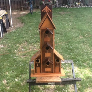 May include: A wooden birdhouse with multiple nesting compartments, painted in shades of brown and featuring a decorative roof design. The birdhouse is mounted on a black metal stand.