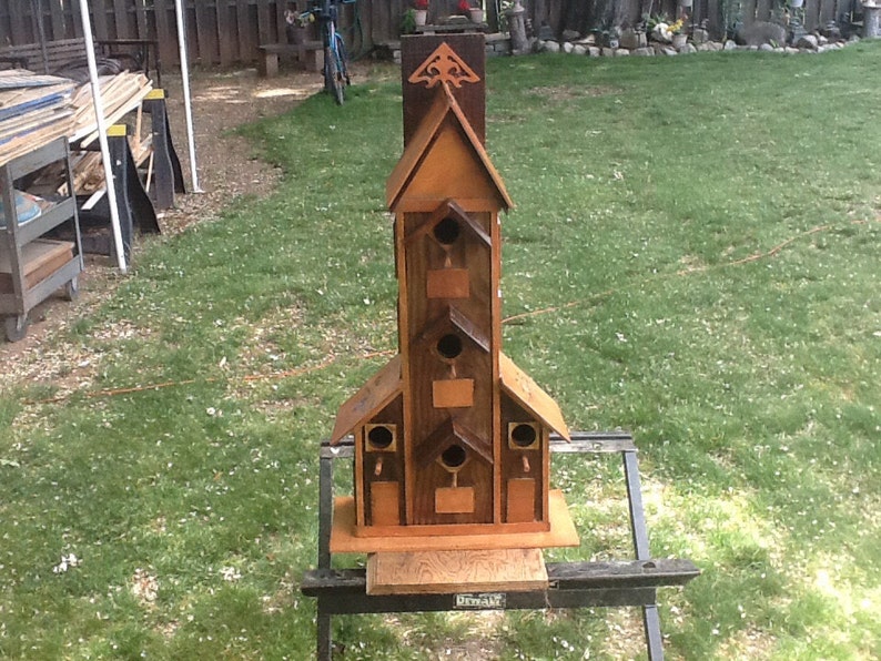May include: A wooden birdhouse with multiple nesting compartments, painted in shades of brown and featuring a decorative roof design. The birdhouse is mounted on a black metal stand.